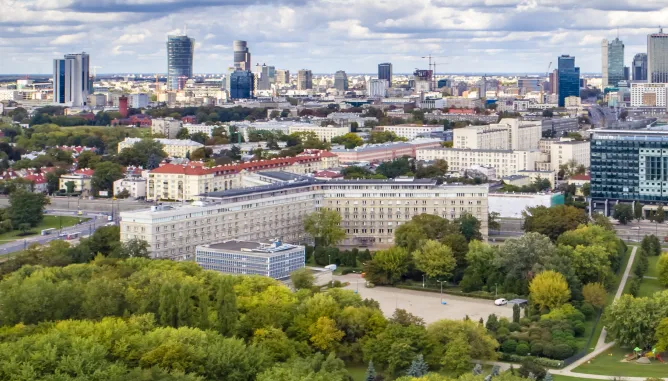 Widok budynku Głównego Urzędu Statystycznego w kształcie litery Y otoczony zielonymi drzewami. W tle panorama współczesnej Warszawy.